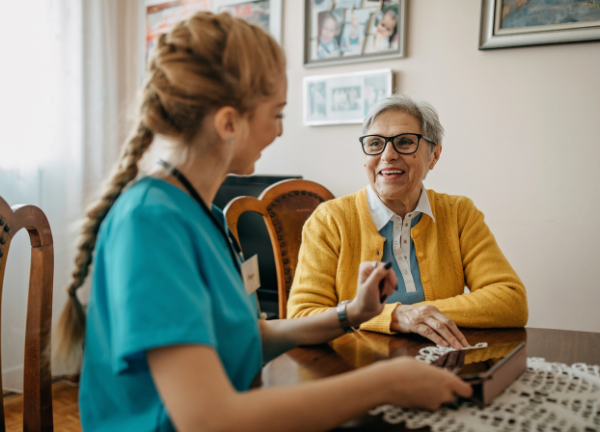 A caregiver in a blue uniform talks with a smiling elderly woman in a yellow cardigan at a dining table.