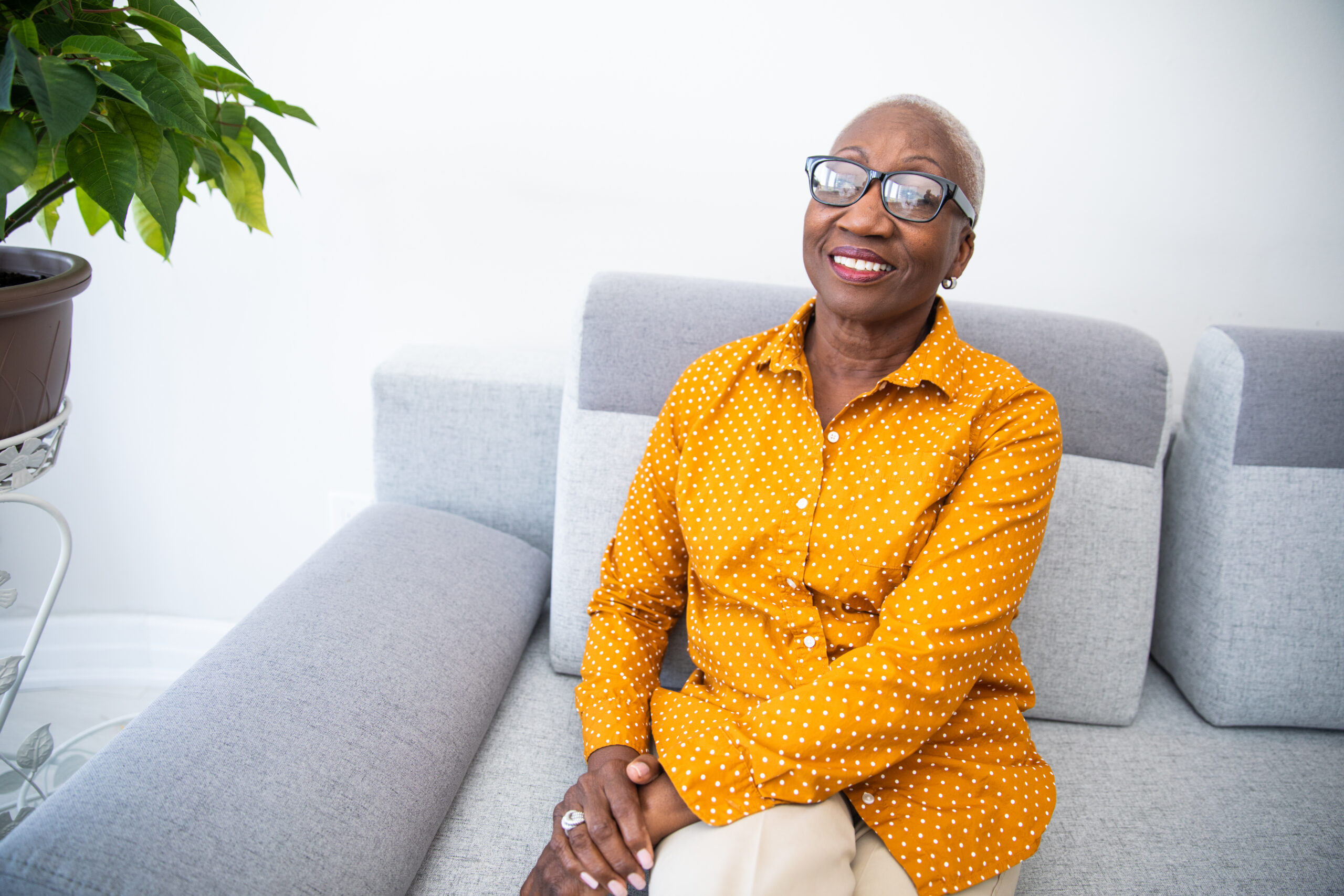 Relaxed Woman A person with glasses smiles while sitting on a gray sofa, wearing a yellow polka dot shirt. A green leafy plant is in the background.