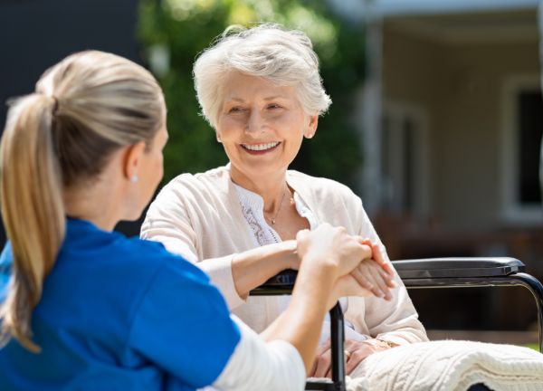 Elderly woman in a wheelchair smiling at a younger woman in a blue uniform.